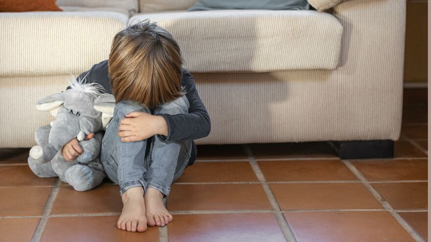 Young child sitting alone on tiled floor hugging knees, holding stuffed elephant, representing scapegoat child concept.
