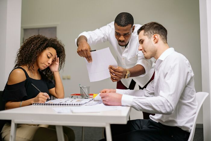 Three professionals at a table. A woman looks distressed while a man shows a document to another man. Workplace incidents and stress.