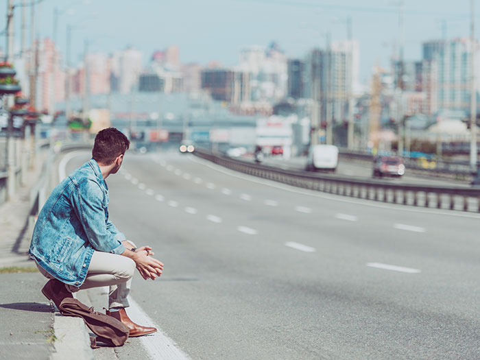 A man crouches by a highway, looking away. His posture suggests waiting or disappointment, reminiscent of horrible first dates.