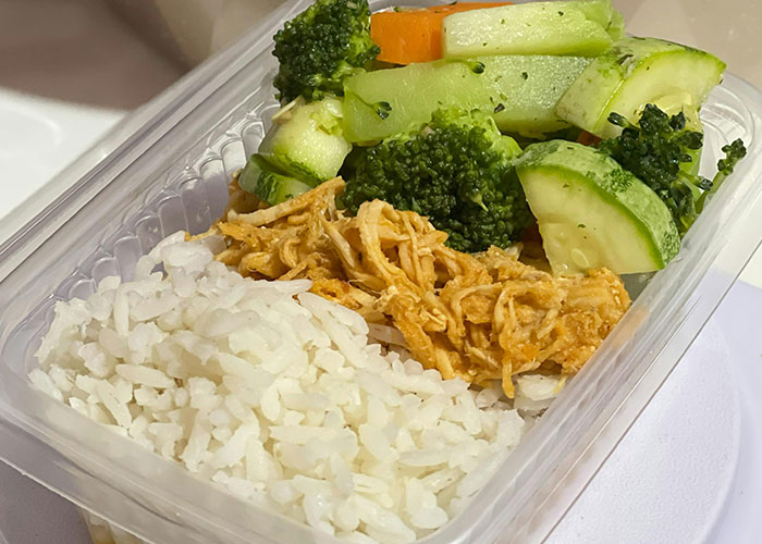 Takeout container with rice, shredded chicken, and mixed steamed vegetables highlighting economic challenges signs.