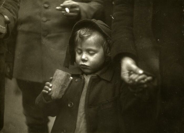 Child holding a piece of bread, wearing a hooded coat, captured in a rare and interesting photo from a past era.