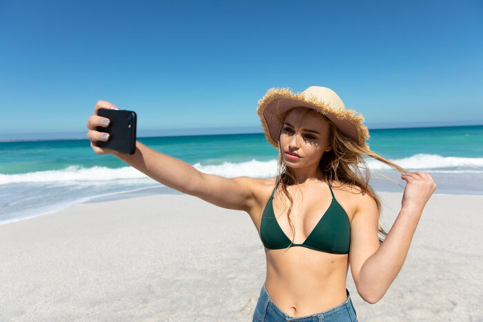 Woman in a sunhat taking a selfie on the beach, capturing a first date moment with red flags and instant regret vibes.