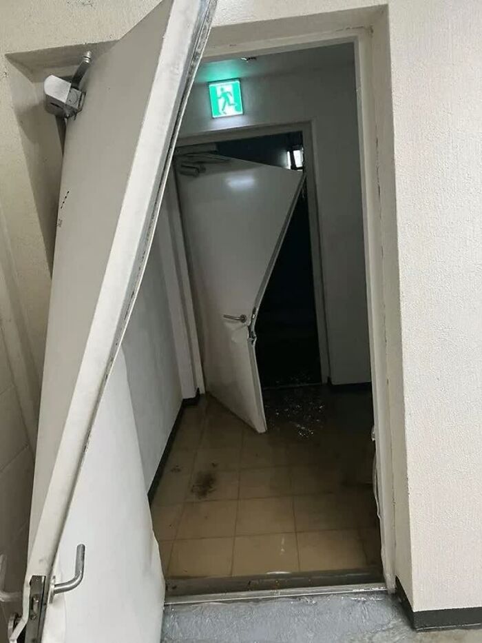 A weird image of two white damaged doors with an exit sign overhead. Water and debris are on the floor.