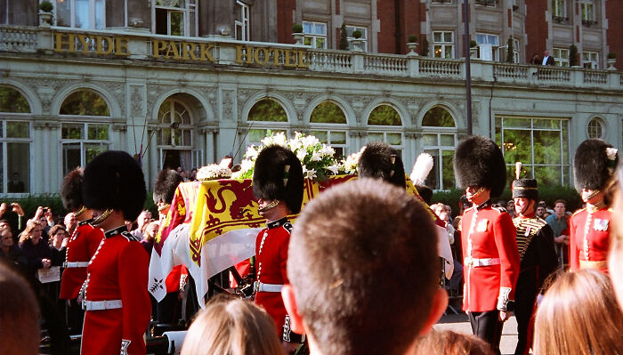 Ceremonial guards in red uniforms carrying a decorated coffin during a public procession outside Hyde Park Hotel.