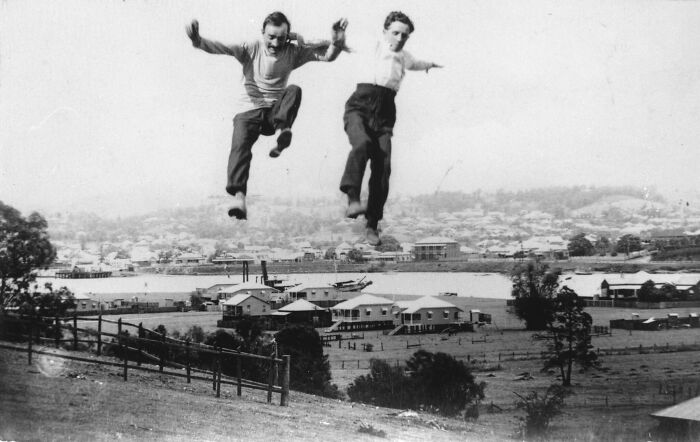 Two men jumping in mid-air over a hill with a historical town and waterfront in the background, rare and interesting photo.