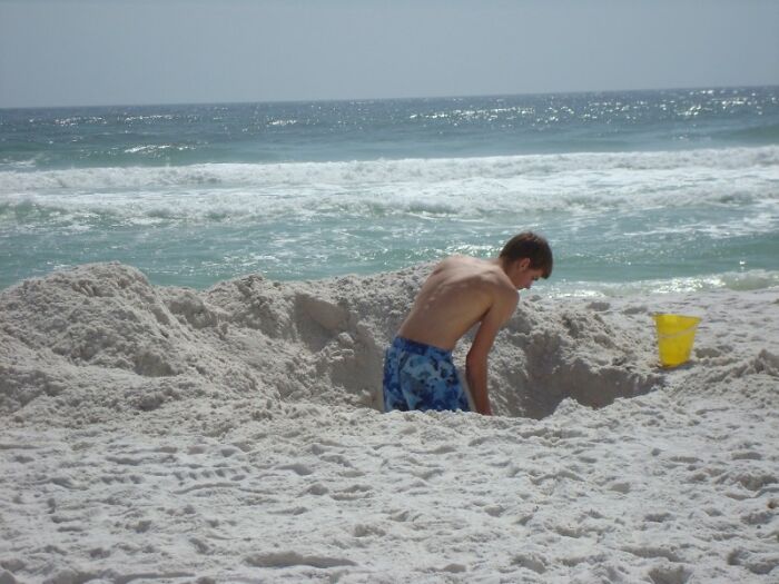 Boy in blue swim trunks digging a large hole in white sand at the beach with ocean waves in the background, terrifying statistics.