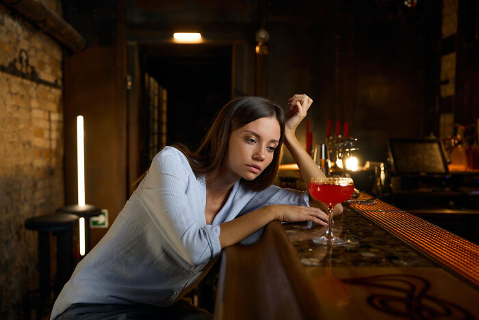 Young woman sitting alone at bar with a red cocktail, appearing disappointed after a first date with red flags and regret.