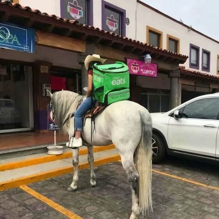 Person wearing a cowboy hat riding a horse with a food delivery backpack outside a storefront in a parking lot.