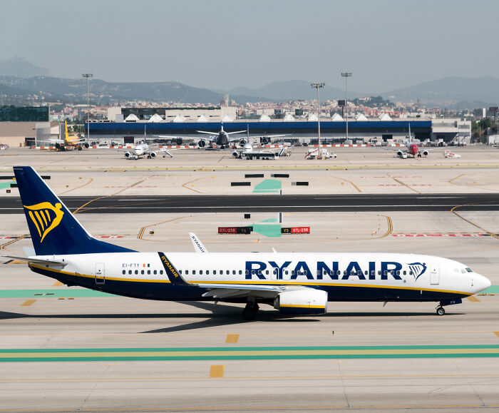 Ryanair airplane taxiing on airport runway with terminal and other planes in the background on a clear day.