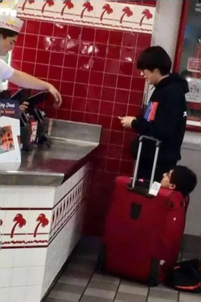 A weird image of a person checking out at a counter, with another person's head sticking out of a red suitcase nearby.