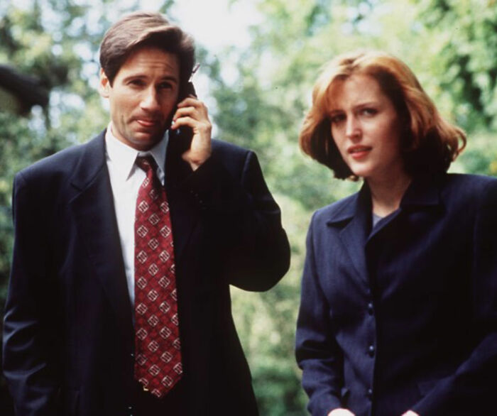 Duas estrelas de TV dos anos 90 de terno do lado de fora, uma ao telefone, ambas parecendo sérias durante uma filmagem em um ambiente arborizado