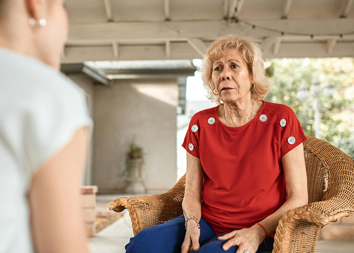 Older woman looking uncomfortable sitting in a chair while a person talks to her inside a house setting.