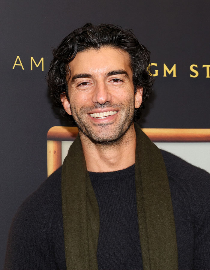 Homem sorridente com cabelo escuro e encaracolado, vestindo um suéter preto e lenço verde em um evento formal, relacionado à batalha legal de Blake Lively.
