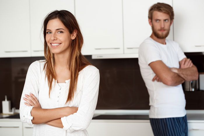 Woman smiling confidently with arms crossed while man looks uncomfortable in a kitchen setting, illustrating wild ways women unsettle men.