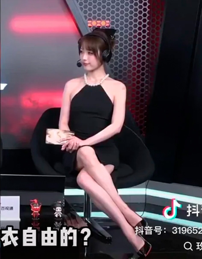 Esports host wearing a black dress and headset, sitting with legs crossed during a live broadcast stage segment. Esports host wearing a black dress and headset, sitting with legs crossed during a live broadcast stage segment.