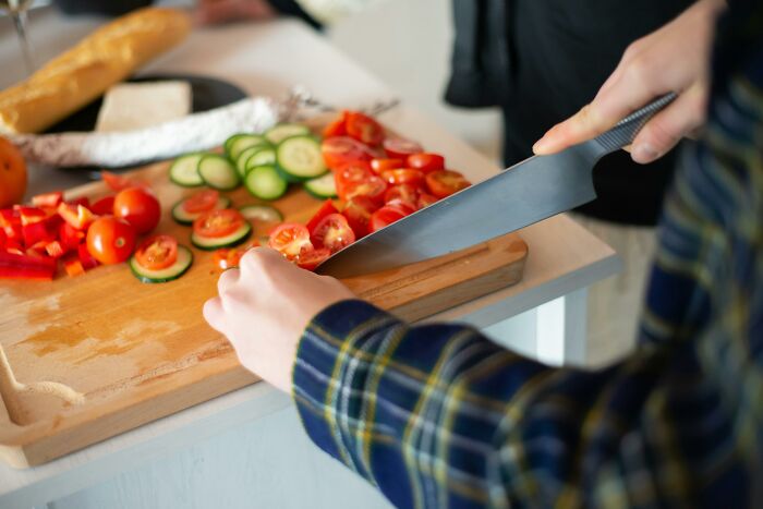 A person in a plaid shirt carefully chopping tomatoes and cucumbers on a wooden board. A reminder of workplace incidents.