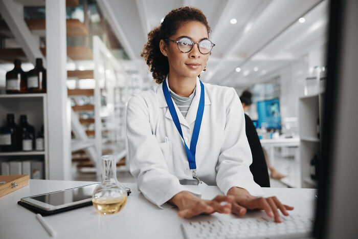 Female medical worker in lab coat and glasses, typing at a computer desk with a flask, pondering difficult experiences.