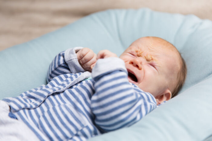 A baby in blue and white striped pajamas cries with eyes squeezed shut, making disturbing sounds on a light blue cushion.
