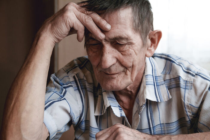 Elderly man with hand to forehead, eyes closed, appearing deeply troubled by thoughts of friendship betrayals.