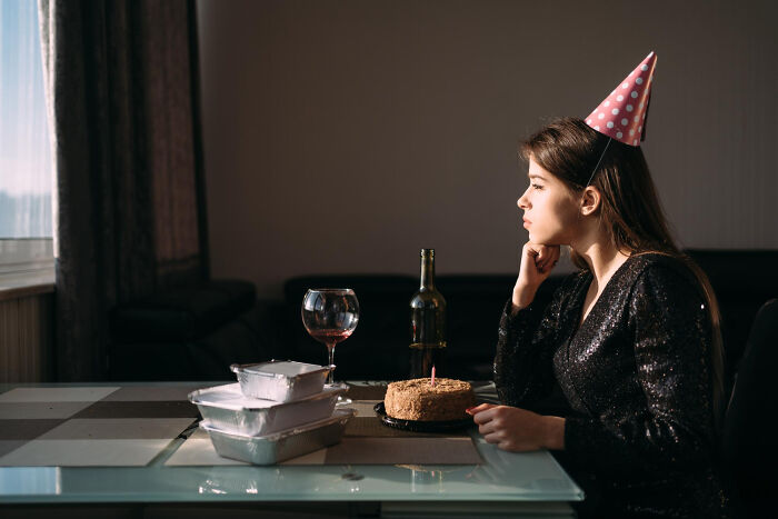 Young woman wearing a party hat, sitting alone with cake and wine, symbolizing good person betrayed and shocked feelings.