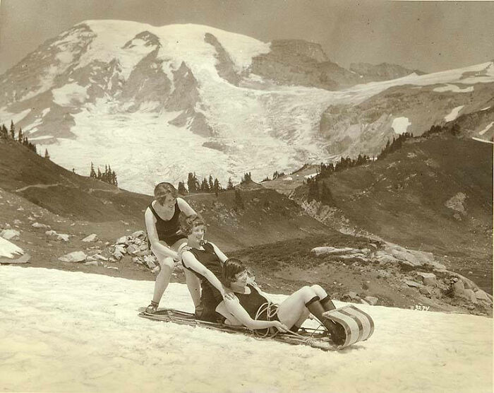 Three women in vintage swimsuits sledding on snow with a mountain backdrop in a rare and interesting historical photo.