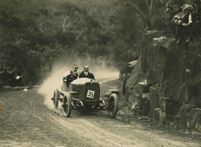 Vintage race car speeding on a dusty road with two men inside, captured in a rare and interesting historic photo.