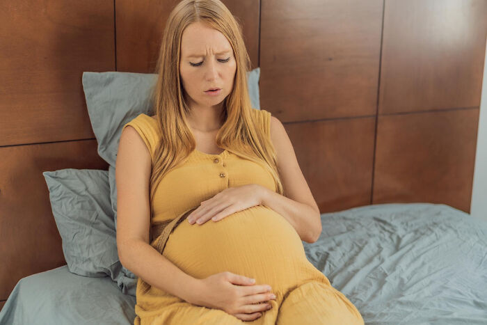 Pregnant woman in yellow dress sitting on bed, holding belly, showing concern about random facts learned about their bodies.
