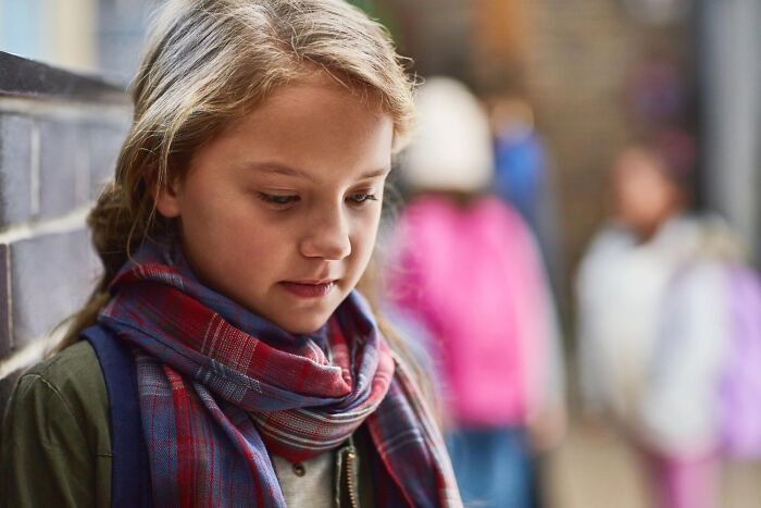 Young girl looking sad near wall while blurred kids in the background, illustrating life dealing with horrible bullies concept.