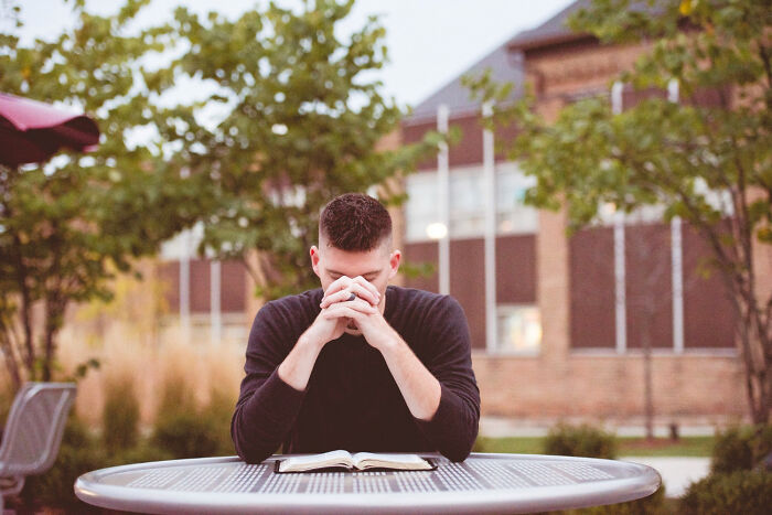 A man with his head down and hands clasped, praying over an open book at an outdoor table. Represents priests and nuns who leave the church.