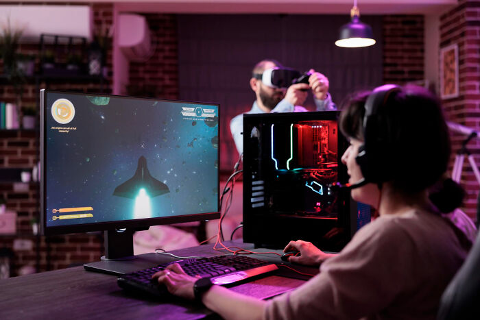 Person playing a computer game with a headset in a dim room while another person uses VR, illustrating random life decisions.
