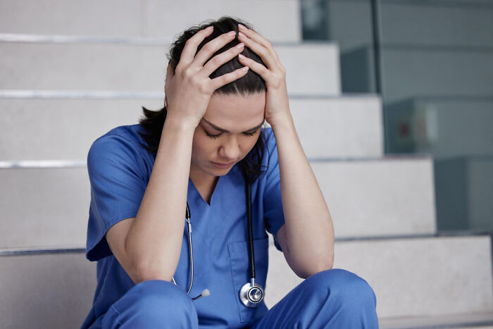 Stressed nurse in blue scrubs sitting on stairs, holding head in hands, reflecting hidden truths about hospitals.