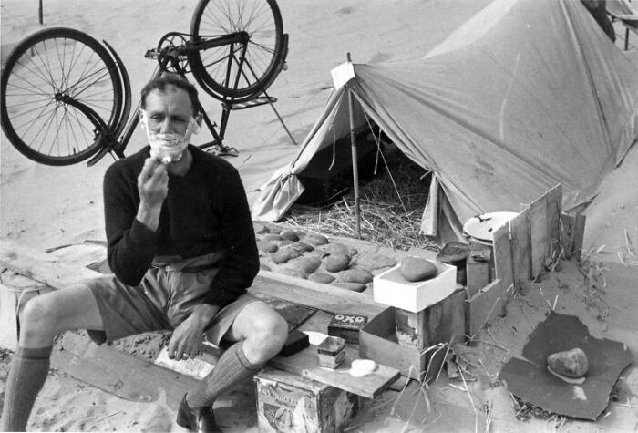 Man shaving outdoors next to a tent with a bicycle hanging, a rare and interesting photo from a time long past.