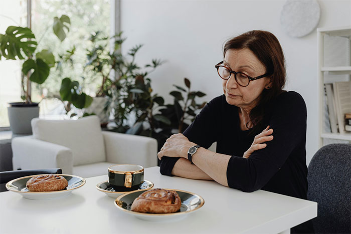 Woman sitting at a table with coffee and pastries, reflecting thoughtfully in a cozy home setting with plants.