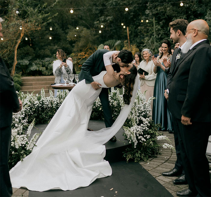 Bride and groom share a kiss at an outdoor wedding ceremony surrounded by guests under string lights and floral decorations