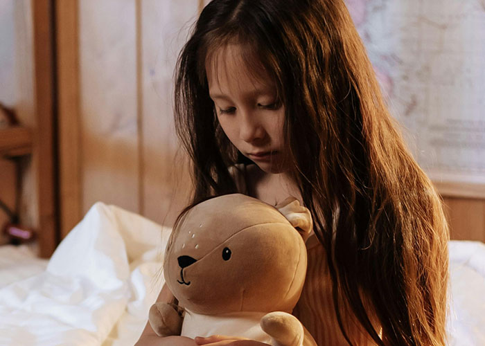 Menina segurando um urso de pelúcia com força, parecendo triste e vulnerável, refletindo que uma mãe não vai acreditar ou defender seu momento de criança.