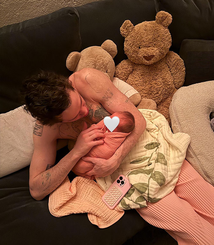 Pete Davidson holding his newborn daughter on a couch surrounded by teddy bears and soft blankets.