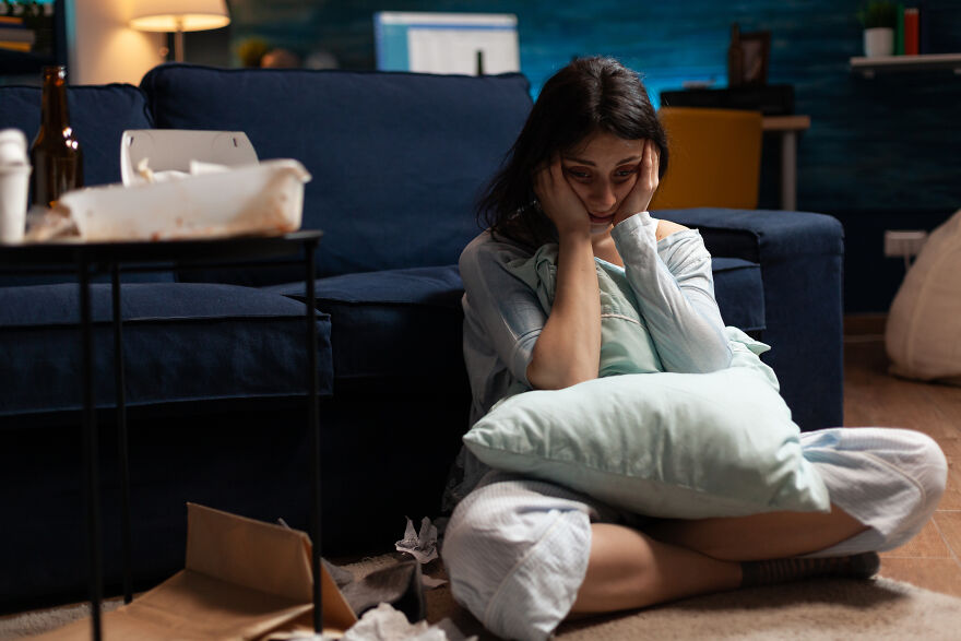 Sad young woman sitting on the floor holding a pillow, illustrating the scapegoat child feeling isolated in family conflict.