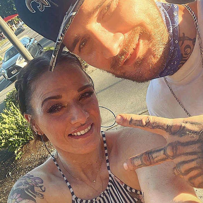 Jovem vista pela última vez colhendo cogumelos, com tatuagens, sorrindo ao ar livre com um homem de boné e bandana.