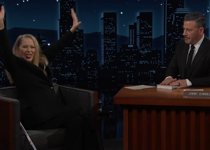 Christina Applegate animatedly raising her arms during a late-night talk show interview with host Jimmy Kimmel.