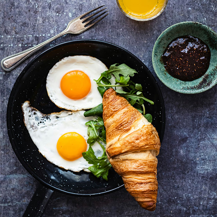Uma frigideira com ovos fritos e verduras com um croissant, sugerindo o potencial de comer ovos no tratamento do Alzheimer.