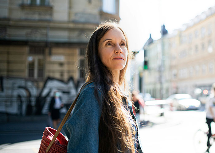 Mulher com longos cabelos castanhos, vestindo uma jaqueta jeans e carregando uma bolsa vermelha, parecendo pensativa em uma rua ensolarada. Os pássaros são sexistas.