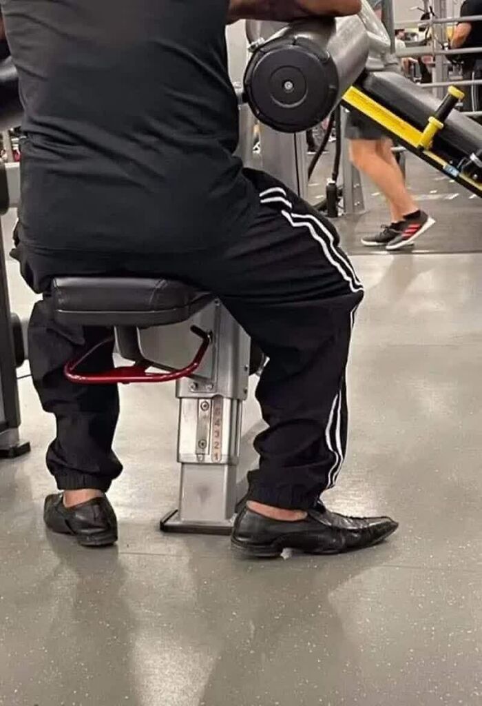 A person in black track pants and dress shoes uses gym equipment, a weird image that lives rent-free in minds.