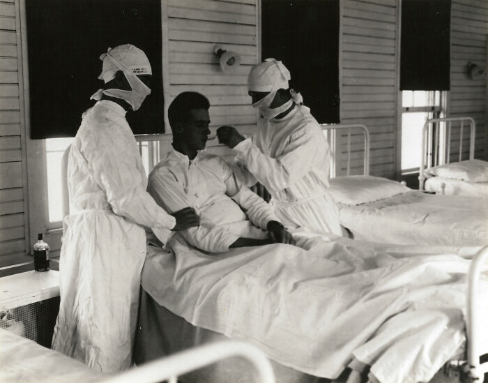 Two medical staff in protective gear treating a patient in a hospital bed in a rare and interesting historical photo.