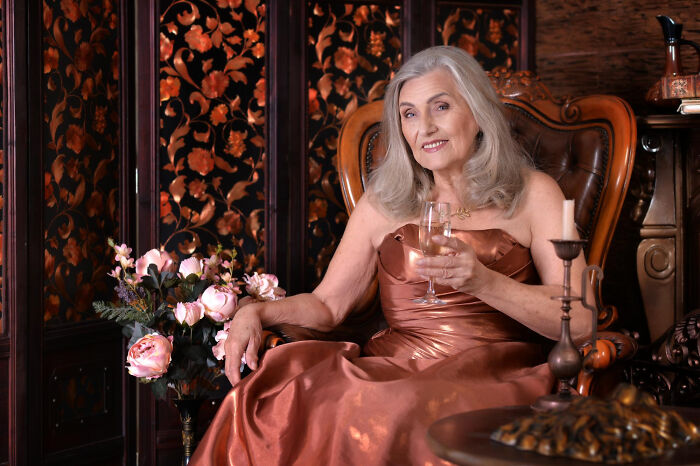 An older woman in a formal dress, holding a champagne flute, representing celebrity relatives and their costs.