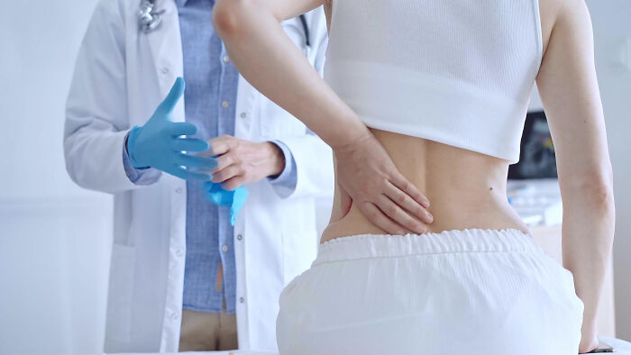 Patient holding lower back in a medical exam room while doctor puts on blue gloves, revealing random facts about bodies.