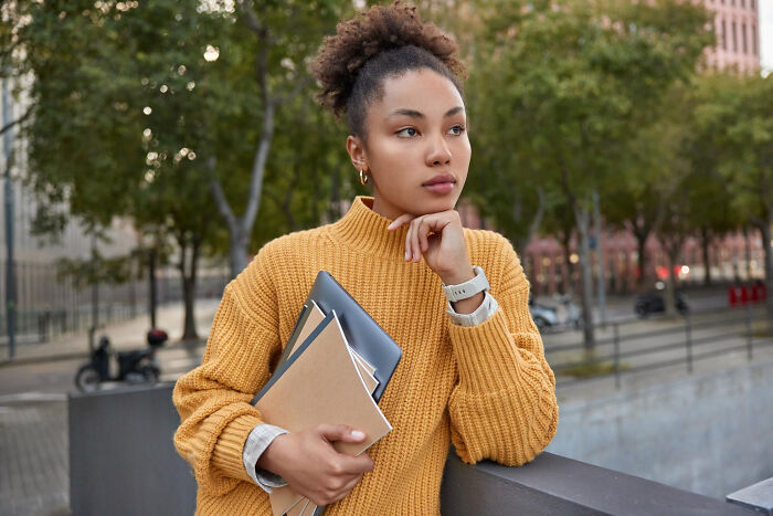 Pensive young woman in yellow sweater holds books, pondering potential friendship betrayals in an urban setting.