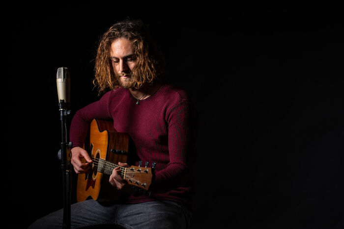 A man with long curly hair in a maroon shirt plays an acoustic guitar into a microphone, reflecting on celebrity relatives.
