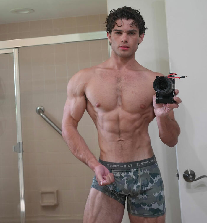 Muscular man taking a mirror selfie in camo underwear, showcasing looksmaxxer physique and clavicular definition indoors.