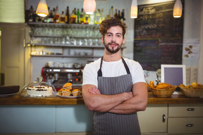 Young barista standing confidently with arms crossed in a cozy cafe, illustrating random life decisions leading to success.