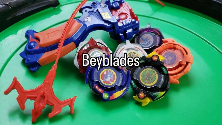 A collection of colorful Beyblades and a launcher on a green arena, evoking nostalgia for growing up in the 2000s.